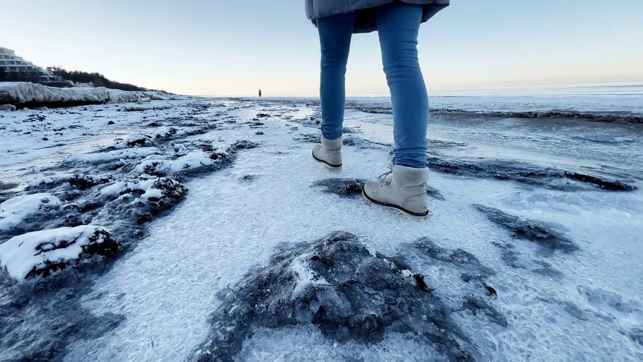 年轻的女人穿着白色的靴子走在冰冻的海滩上踩在冰上，使它裂开视频下载