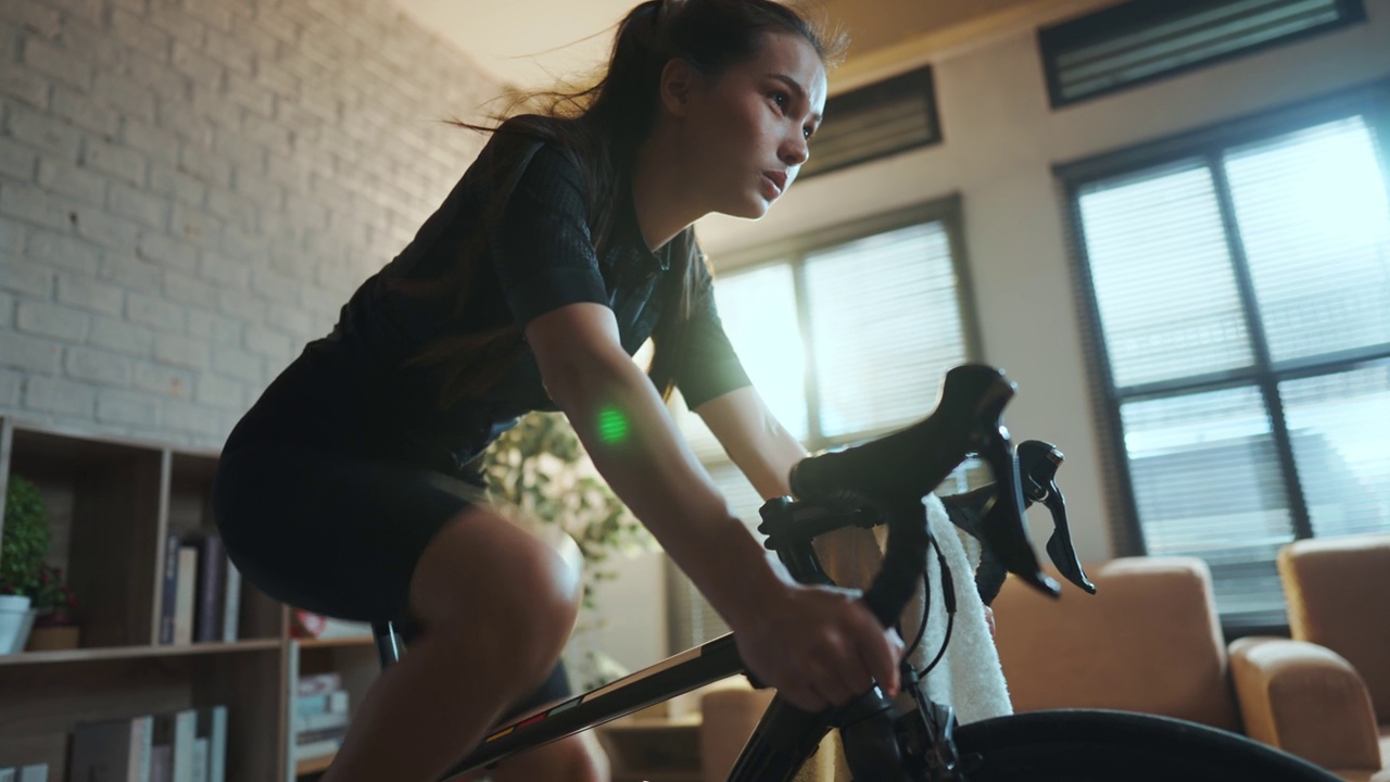 亚洲女人骑自行车。她正在房子里锻炼。通过骑自行车上的教练和玩在线自行车游戏视频素材