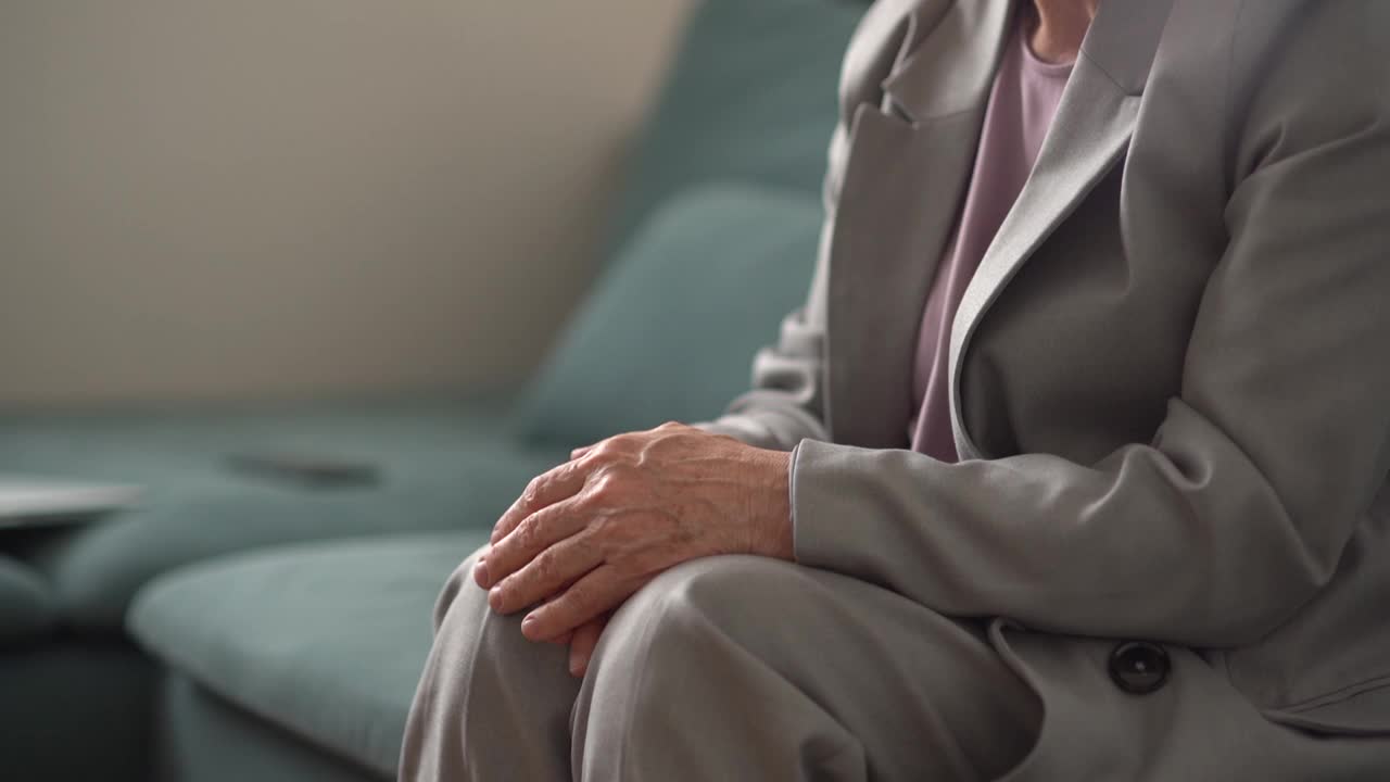 一位悲伤的老妇人正坐在家里的沙发上，她的丈夫走过来，握住了她的手。一对老年夫妇的手的特写，支持和温柔的概念视频下载