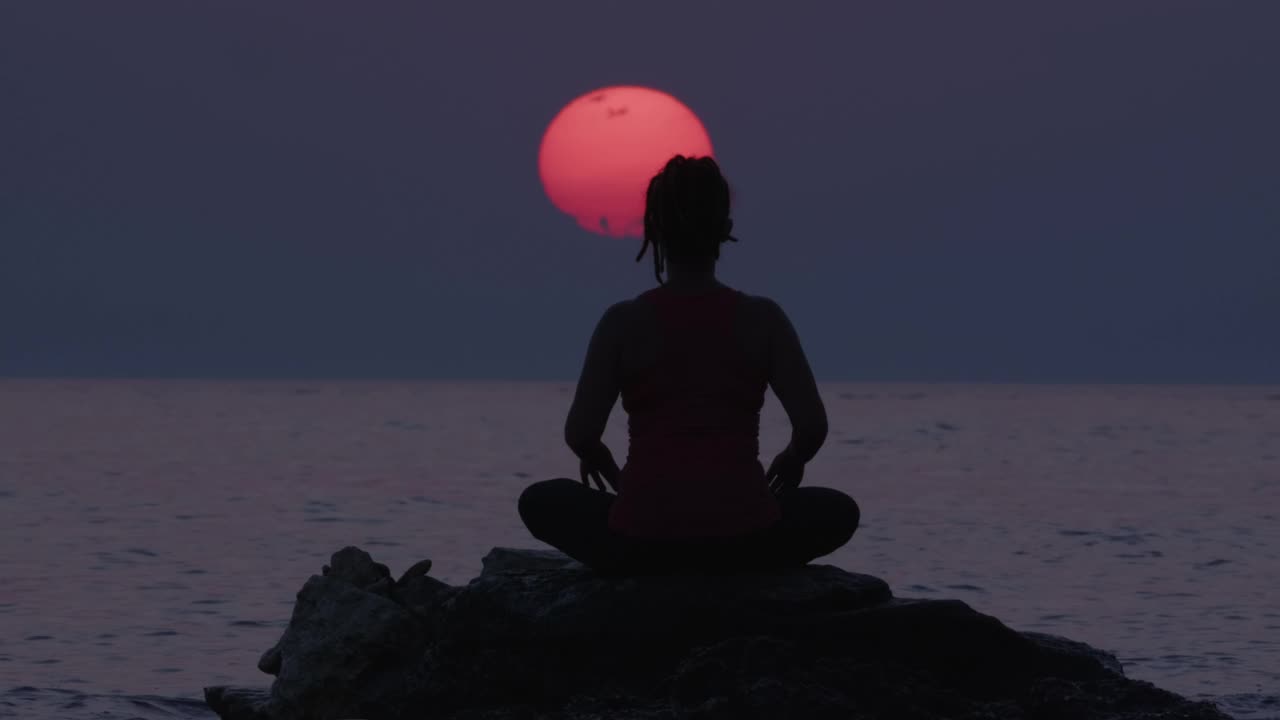 在令人惊叹的日落天空和大太阳期间，坐在海水中的石头上冥想的女人的剪影。治疗身体，心理和精神状态的人。视频下载