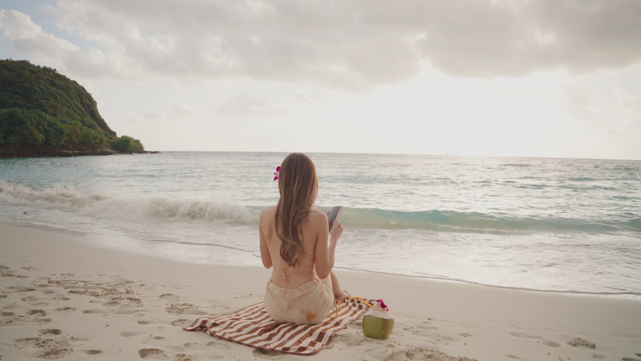亚洲年轻女子穿着泳衣，喝着椰子汁，坐在沙滩上看书。视频下载