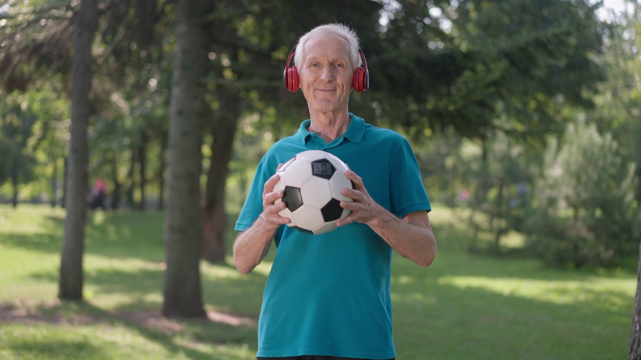 戴着耳机的快乐老人在夏日公园里与足球摆姿势。积极的白人男性退休人员看着相机微笑享受春天户外运动的肖像。长寿的概念。视频下载