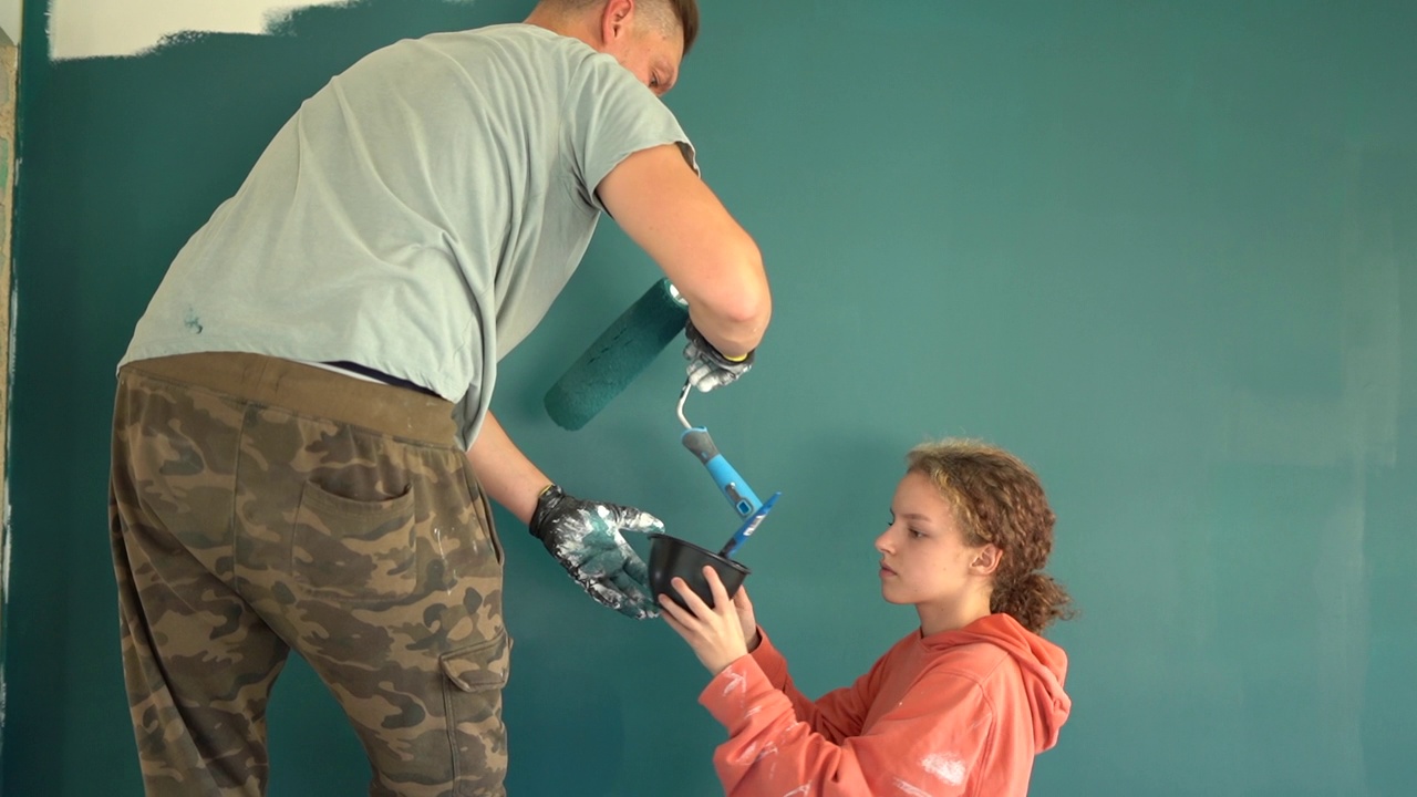 一名男子站在活梯上用蓝色颜料涂墙。他十几岁的女儿帮助他。一个幸福的家庭在修理新公寓视频下载