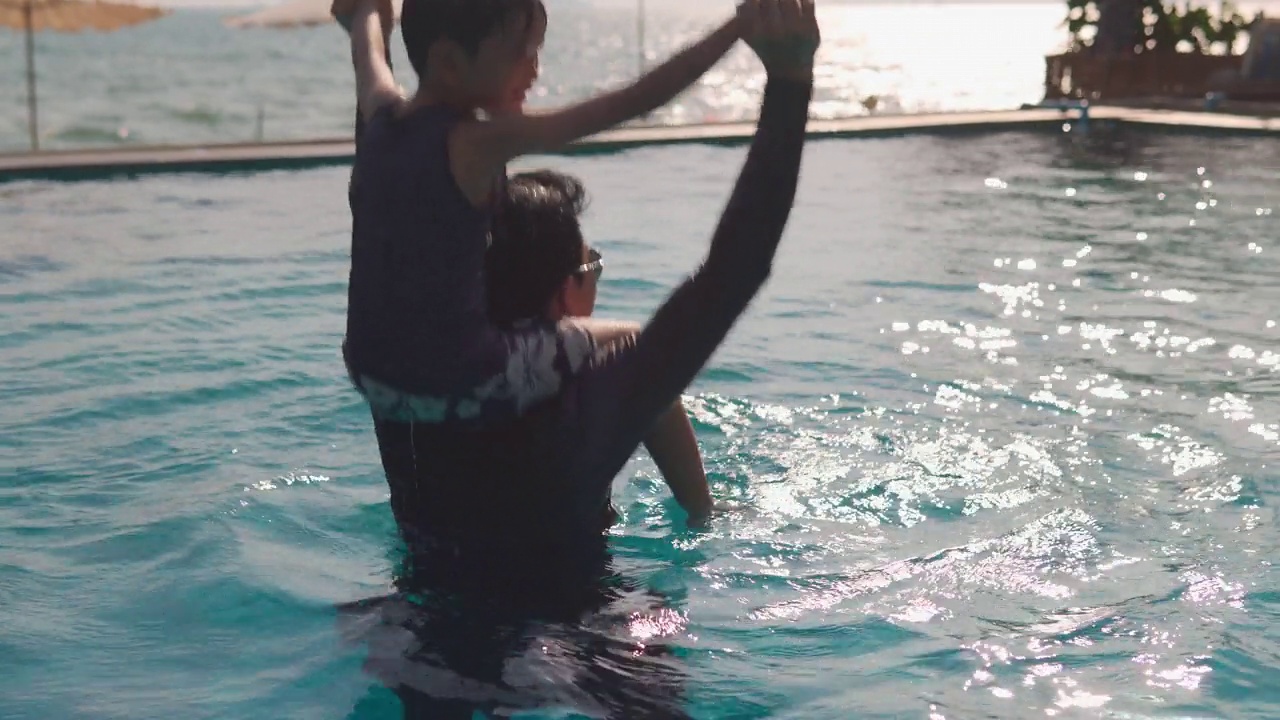 快乐的亚洲家庭:亚洲男孩骑在爸爸的脖子上，爸爸和男孩一起在游泳池里玩耍视频素材