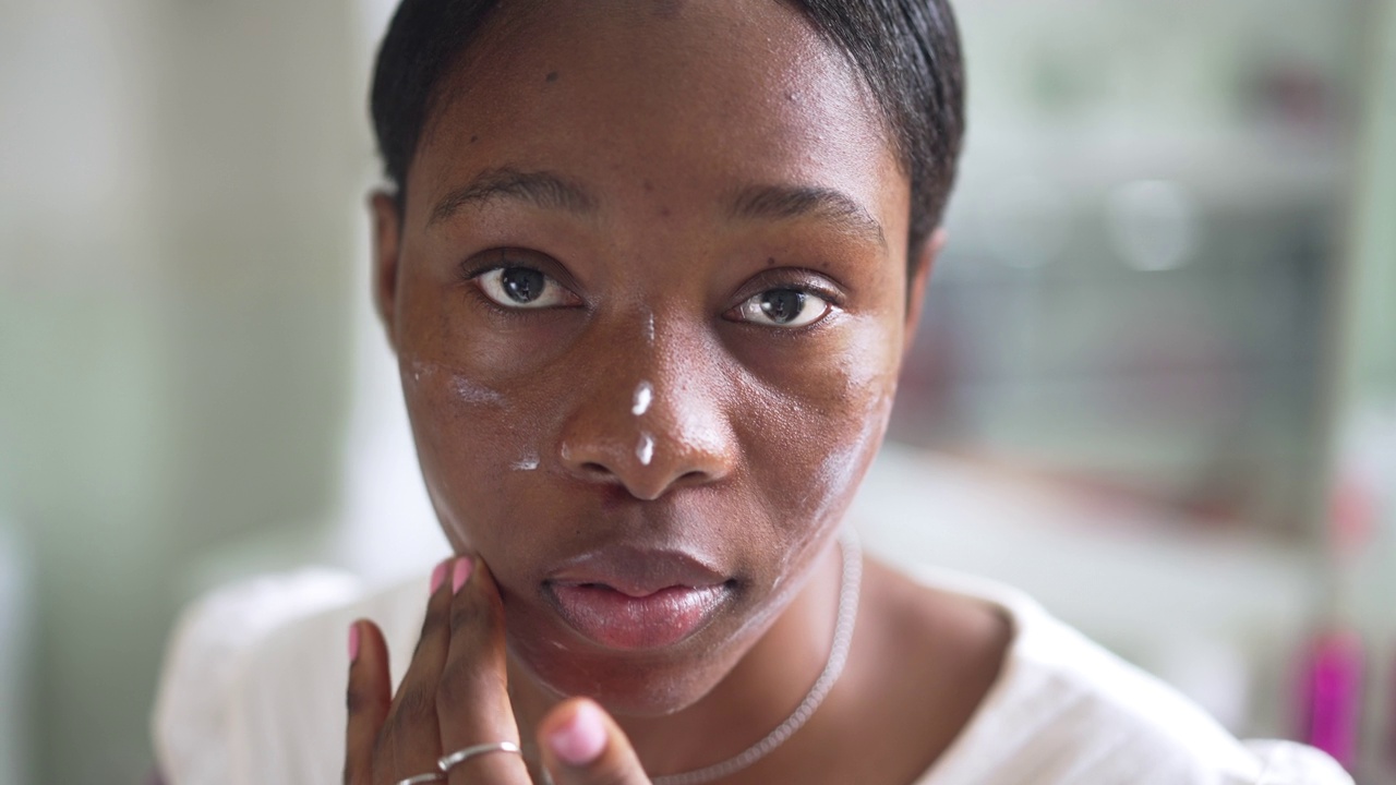 非洲裔美国年轻女性的头部照片，她正在脸上涂抹保湿霜，看着镜头。特写肖像的美丽专注的女士摆姿势照顾皮肤在浴室在家里。缓慢的运动。视频素材