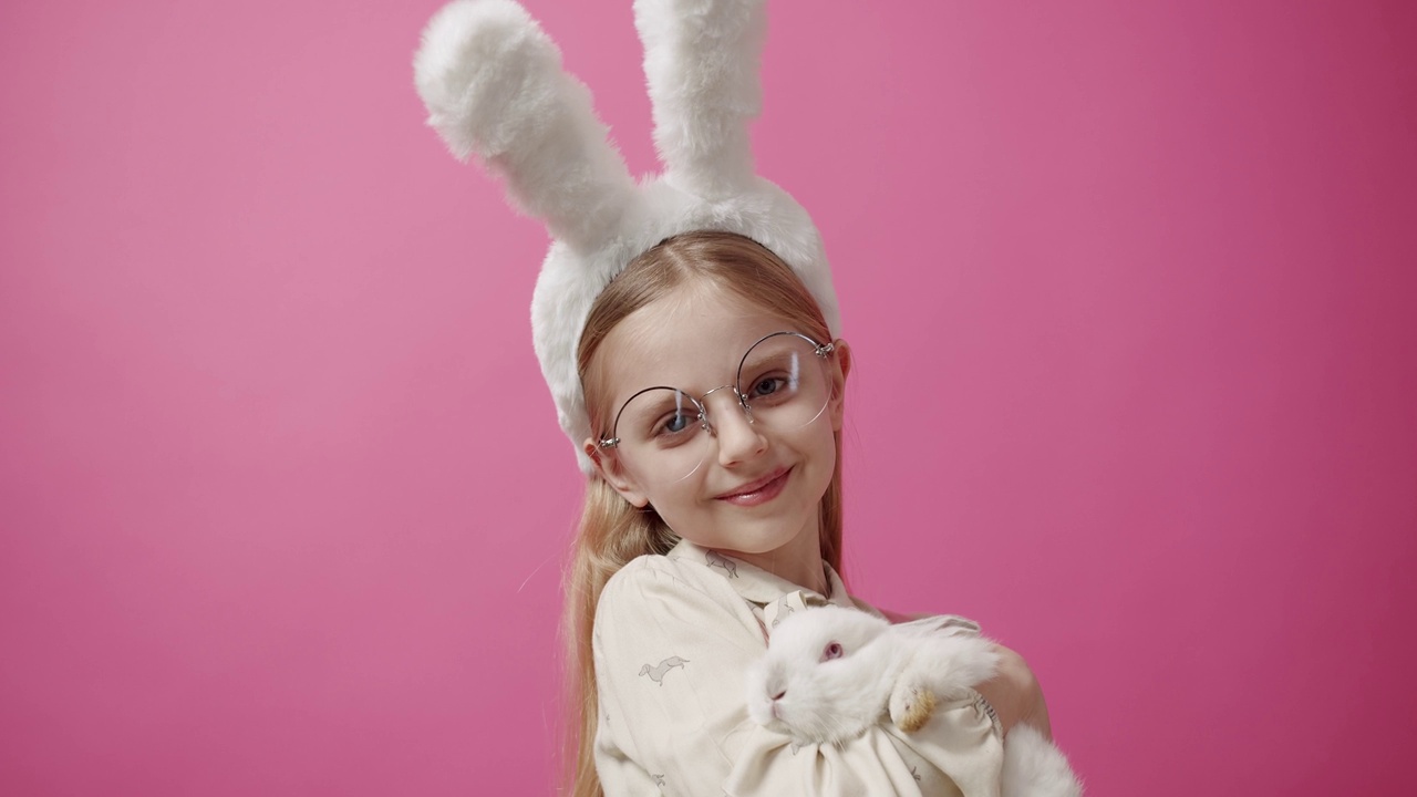 小女孩戴着兔耳朵，手里拿着一只兔子，摆姿势微笑。横幅文本视频下载