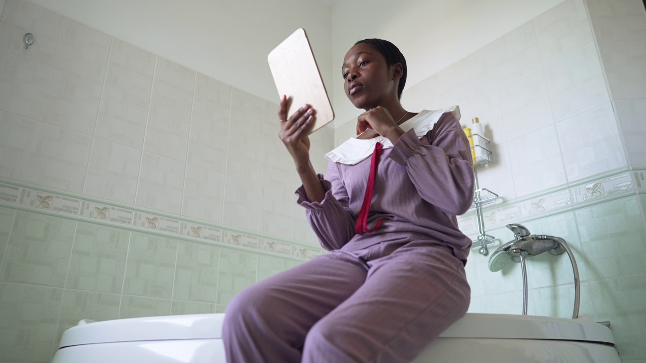 一名年轻的非洲裔美国妇女坐在浴室的浴缸里看着镜子。聚焦在家中的千禧一代女士的肖像。日常美容和生活方式。视频素材