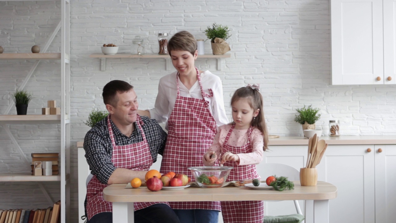 妈妈，爸爸和女儿在厨房做饭。幸福家庭的概念。一个男人，一个年轻女人和一个可爱的小女儿在一起做沙拉。视频下载