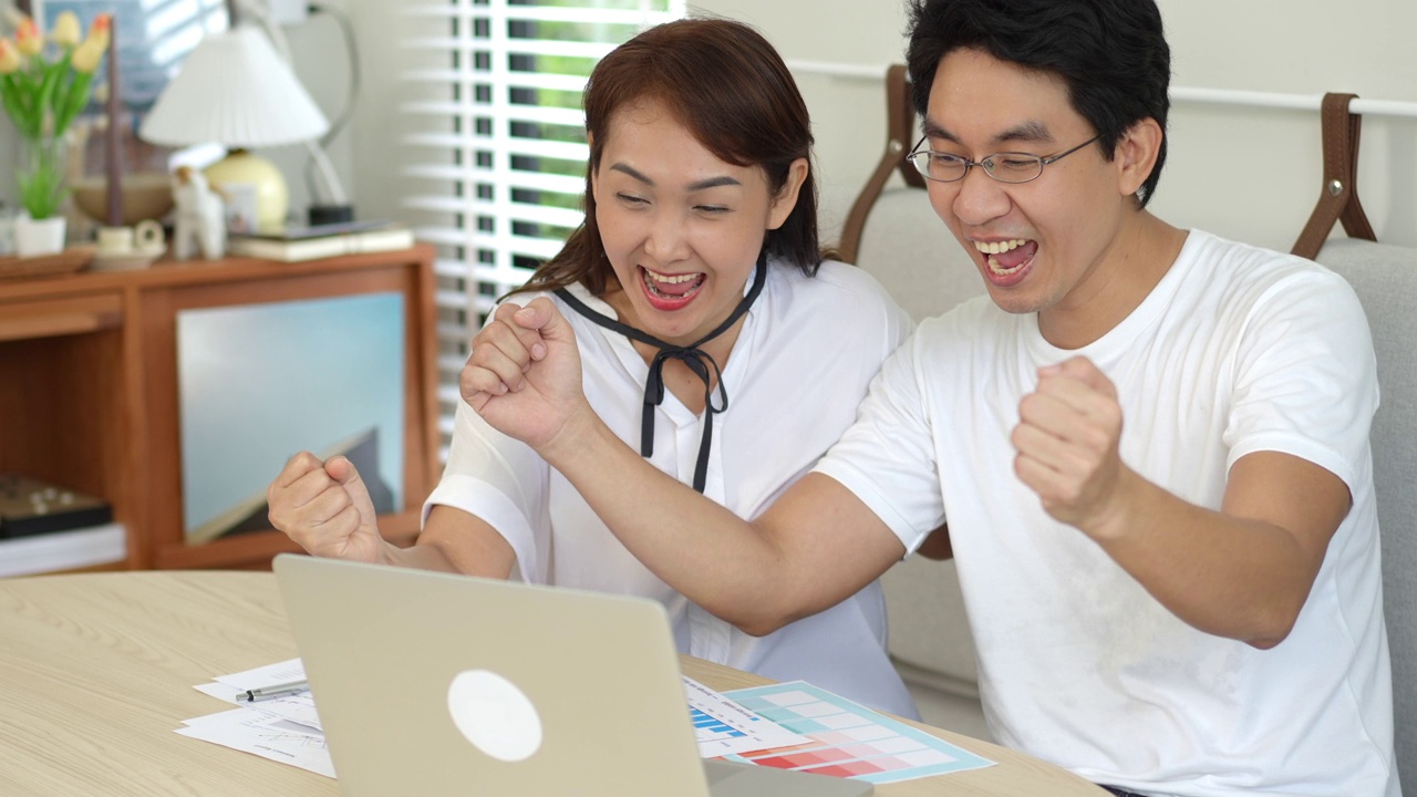 成功的亚洲夫妇夫妇兴奋的好消息从网上笔记本电脑在家里，幸福的夫妻高兴与金融投资的好消息。视频下载