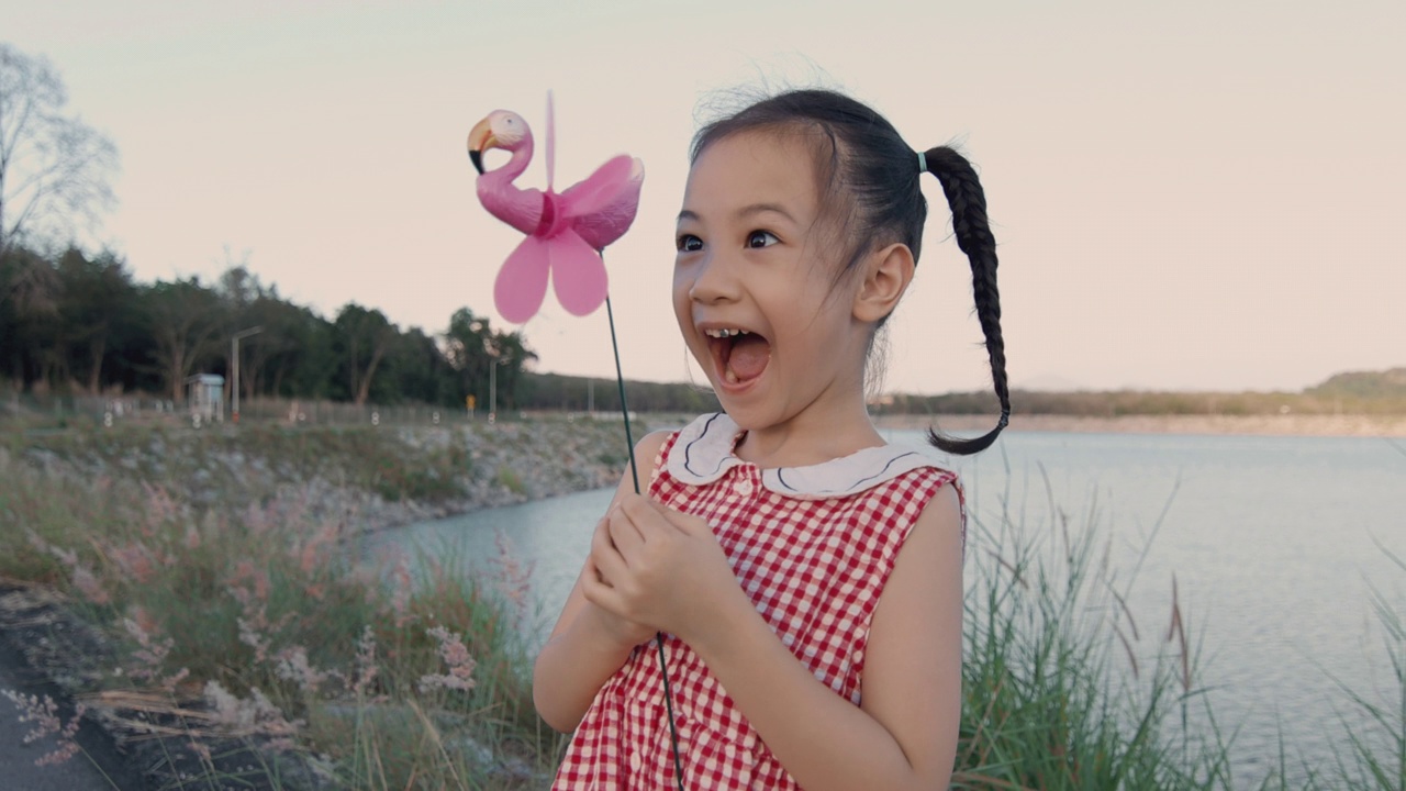 可爱的亚洲女孩在公园玩风车美丽的天空和湖在夏天的时间。以愉悦、快乐、愉悦的表情表现休闲户外活动。视频下载