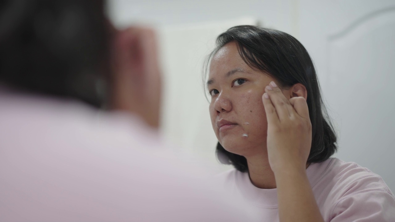 亚洲妇女看着镜子中自己的脸，在使用面霜治疗视频下载