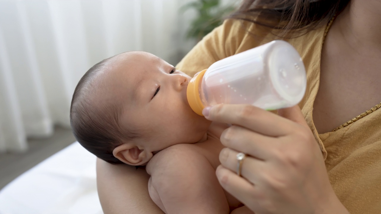 4k，一个两个月大的亚洲新生儿正在睡觉，吮吸着放在妈妈腿上的奶瓶，妈妈的手握着奶瓶。视频素材