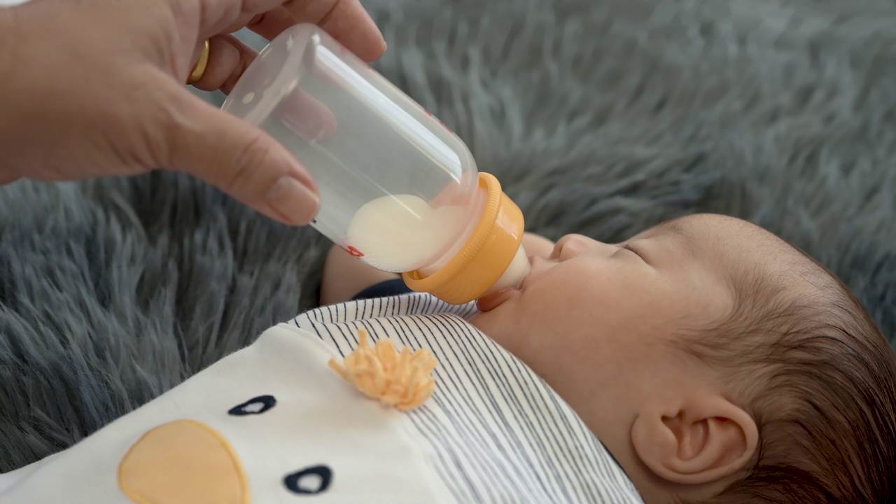 4k，一个2个月大的亚洲新生儿在她妈妈喂的奶瓶里吮吸着奶，睡在灰色的地毯上。视频素材