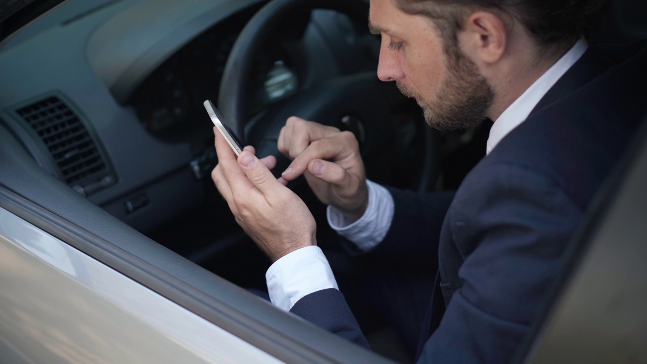 高视角视角，年轻男子坐在驾驶座上，用智能手机发短信，启动GPS导航应用。帅气的白人司机开着智能手机。现代科技和便利。视频下载