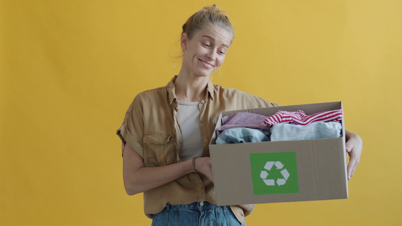一名年轻女子拿着装着旧衣服的纸板箱在黄色背景上进行回收视频下载