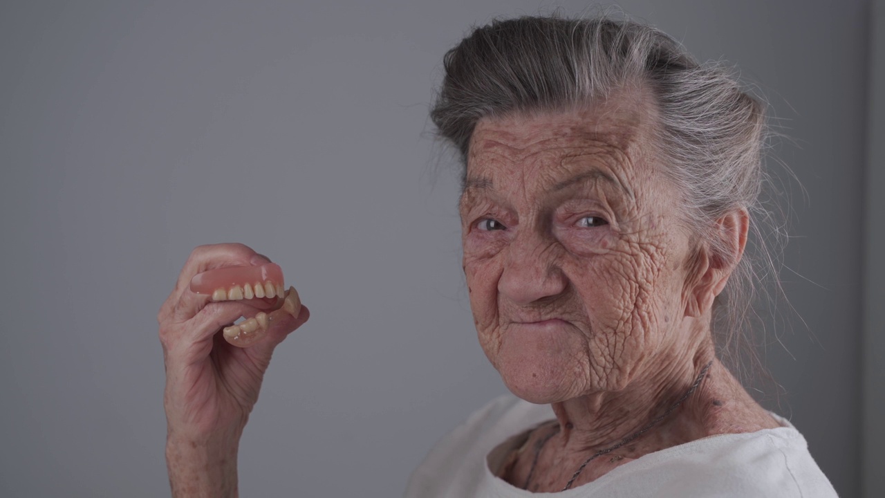 在灰色背景的演播室里，满意的90岁灰发老年妇女手里拿着义齿。没有牙齿的老年妇女微笑着拿着假牙视频下载