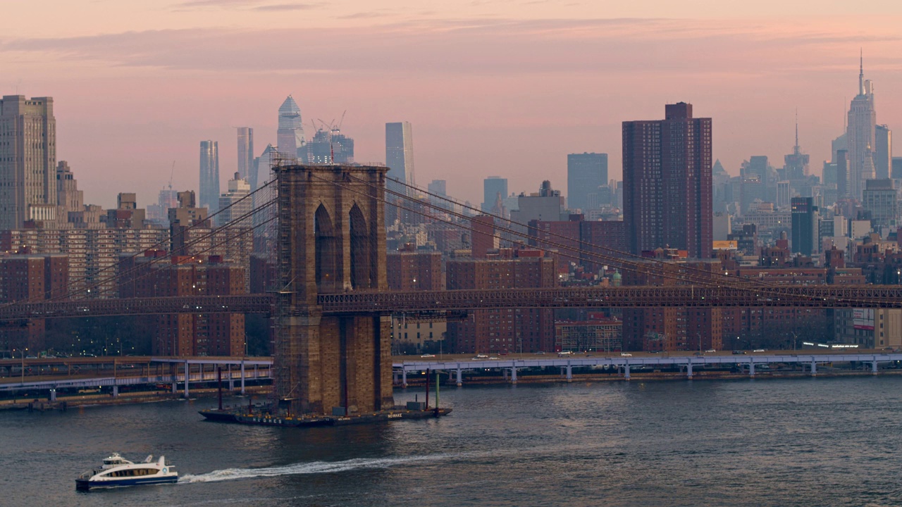 在清晨的黎明时分，一艘渡船在布鲁克林大桥下从空中俯瞰，背景是曼哈顿天际线。无人机视频与平移摄像机运动。视频素材