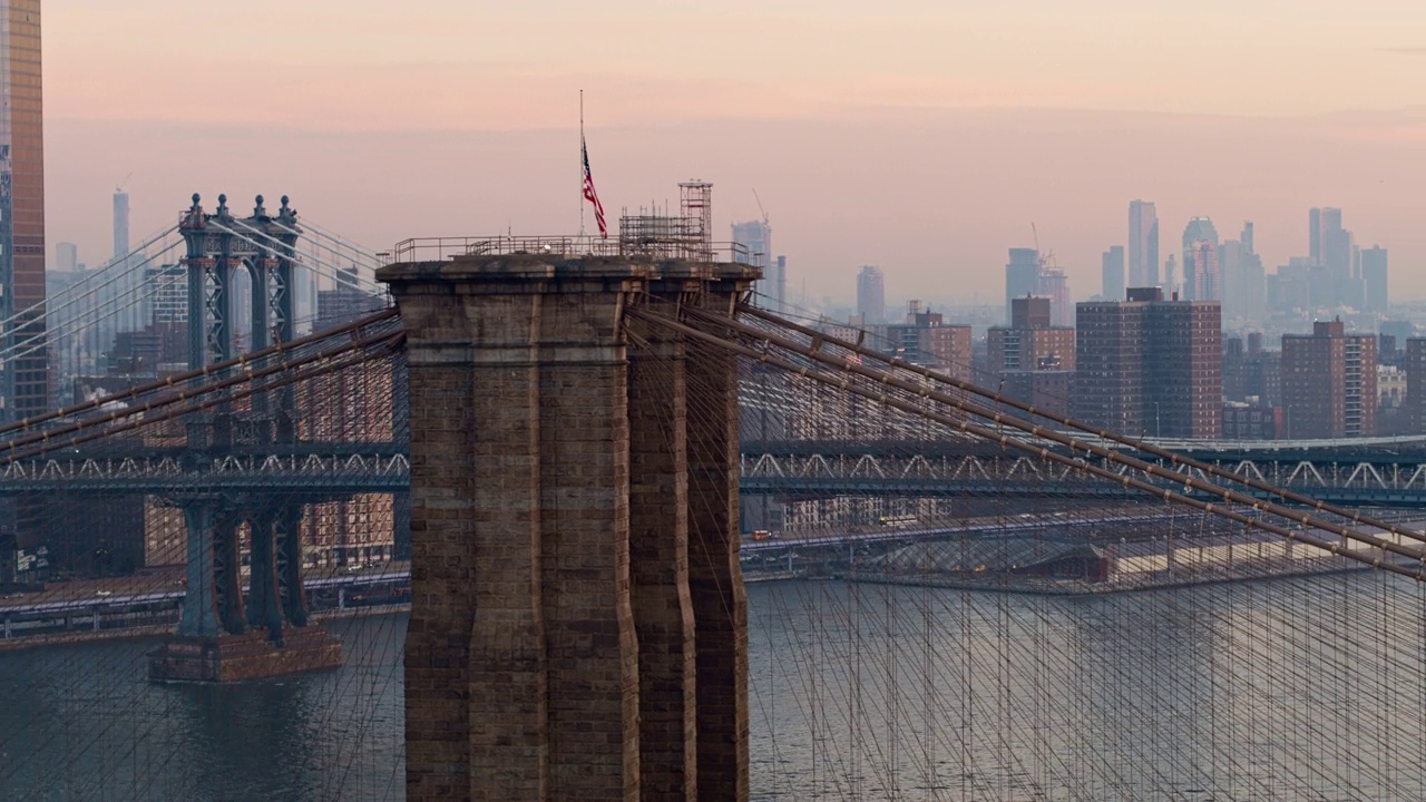 在清晨的黎明时分，俯瞰布鲁克林大桥塔顶飘扬的美国国旗，遥望曼哈顿天际线。无人机视频与平移摄像机运动。视频素材