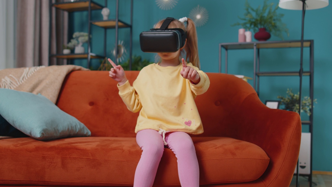 蹒跚学步的小女孩坐在家里的沙发上，使用虚拟现实头盔app玩模拟游戏视频下载