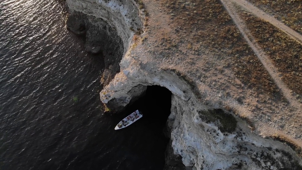 鸟瞰图，一艘摩托艇载着游客在夜晚驶出海岸上的岩石洞穴视频下载