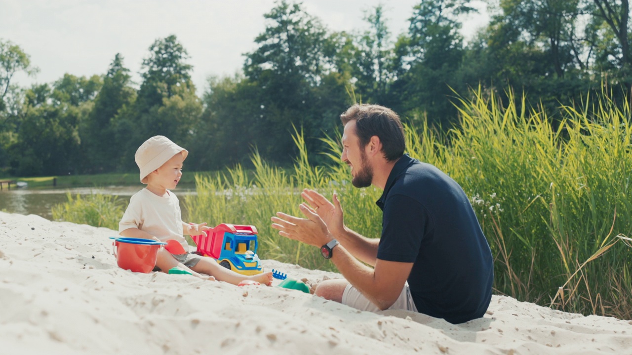 一个爸爸跑到一个戴着帽子的小男孩身边，他在湖边的沙滩上玩，躺在儿子身边，和他说话，他们击掌，孩子伸手去拿铲子视频下载
