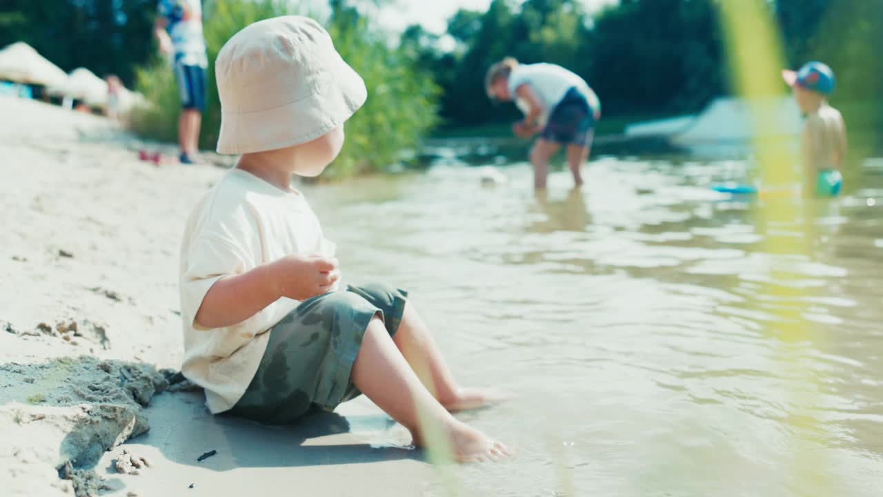 一个戴着帽子的小男孩，穿着短裤和t恤，坐在湖边，看着其他在水里洗澡的孩子，他的拳头在湖面上拍打着，四处飞溅视频下载