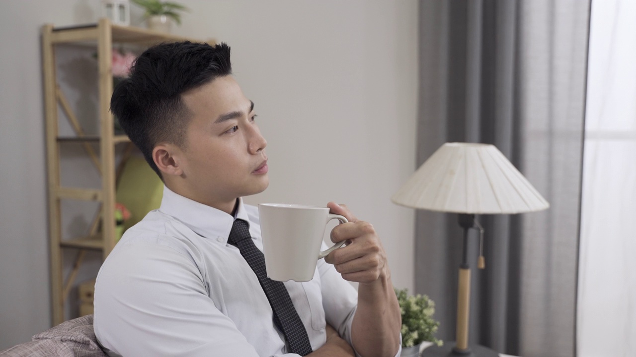 肖像英俊的亚洲商人凝视着远方，抱臂思考，在家里的现代客厅里喝着早。视频下载