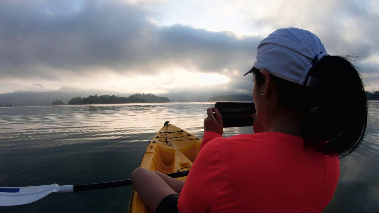 亚洲女子皮划艇在热带大湖和使用手机拍摄日出照片视频下载