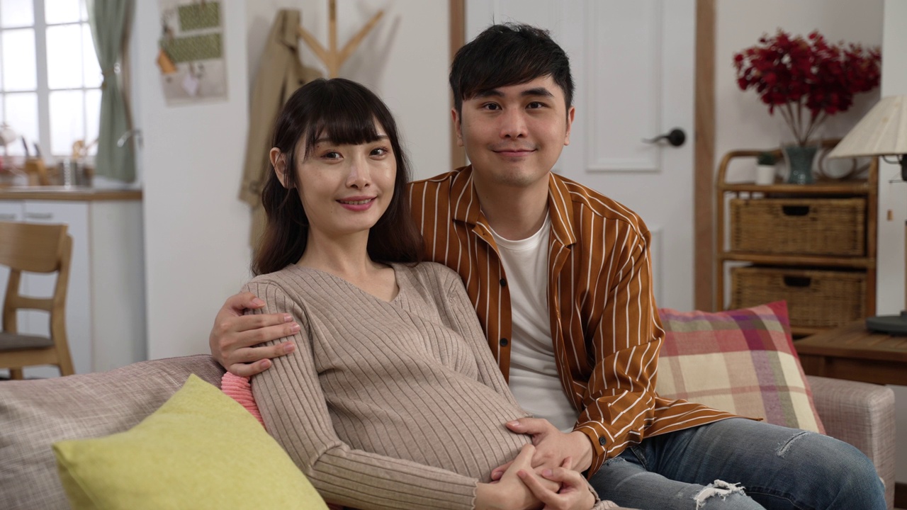 这是一对幸福的亚洲夫妇期待着孩子，在家里客厅对着摄像机微笑的照片视频素材