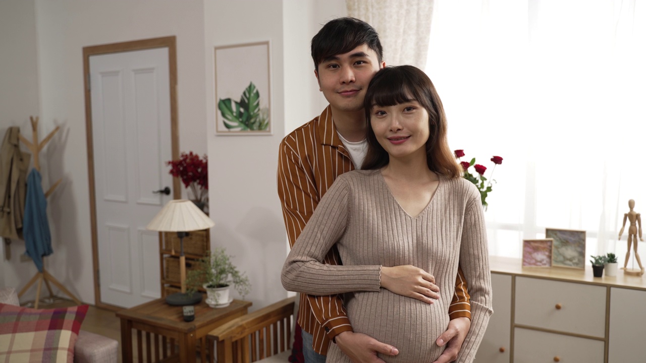 年轻幸福的亚洲夫妇期待一个孩子的肖像，看着相机在家里。丈夫从背后拥抱妻子视频素材