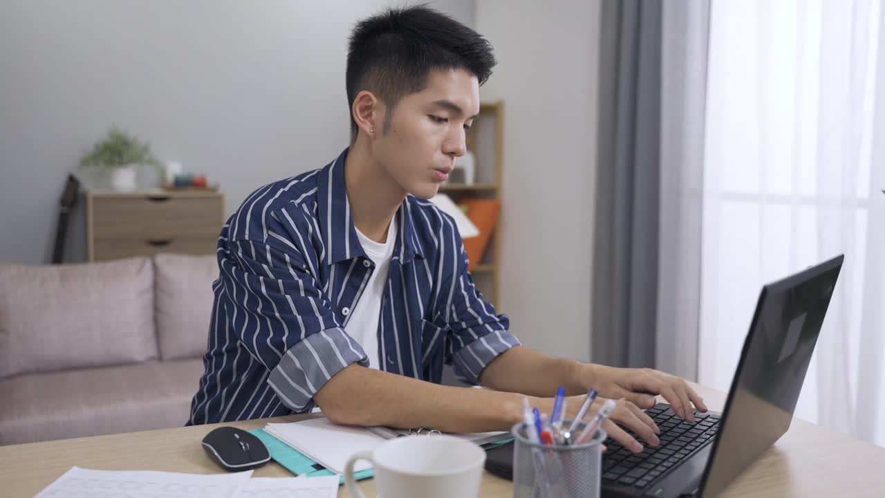 一个在家工作的亚洲人一边在客厅的电脑上打财务报告，一边看报纸。视频素材