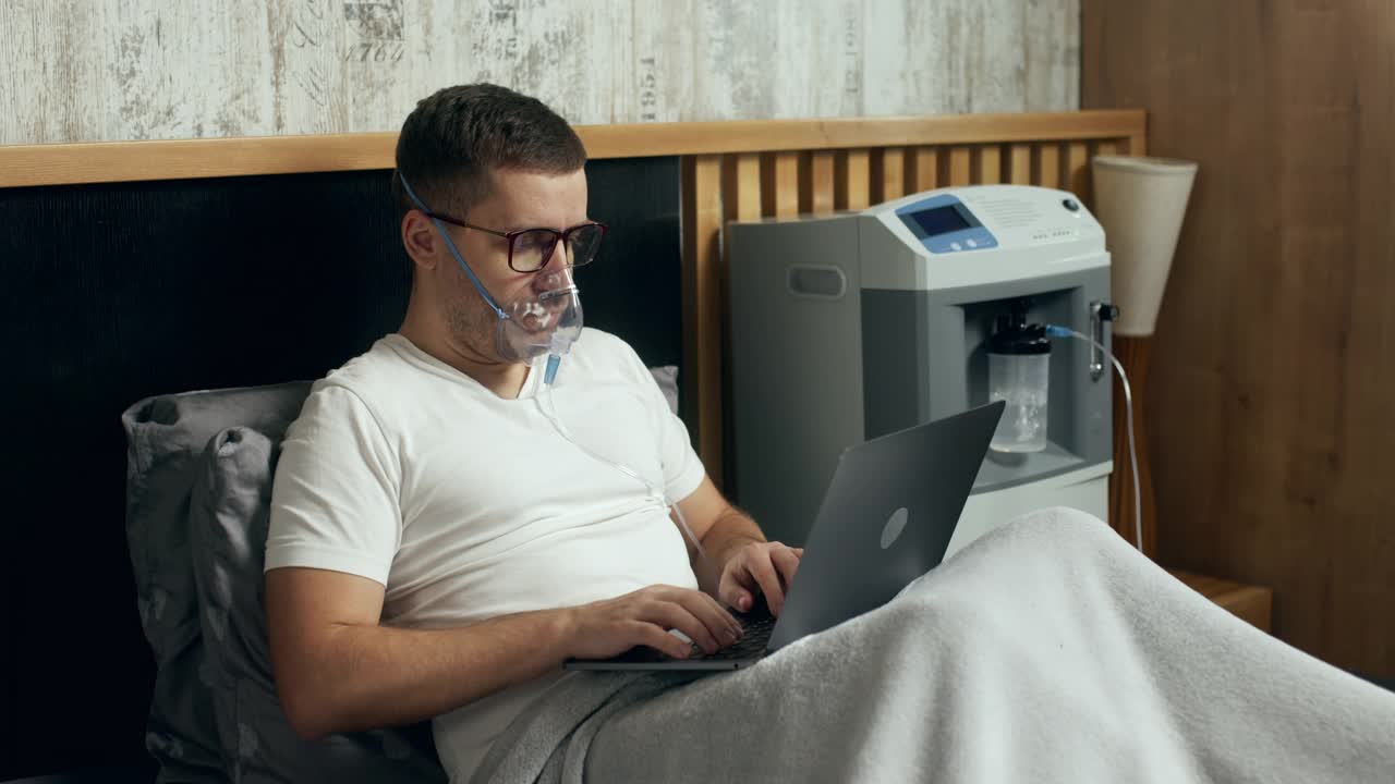 一个生病的年轻人的肖像使用氧气面罩和氧气浓缩器在家里治疗和在笔记本电脑上工作。视频下载