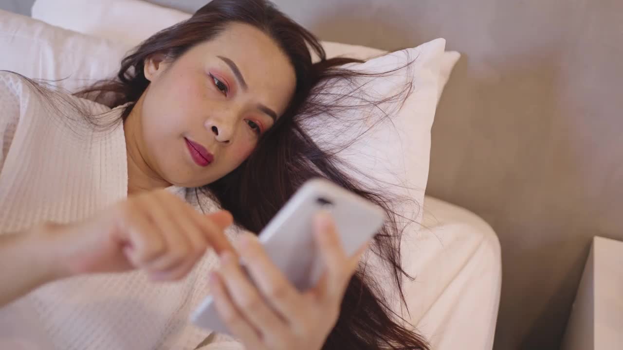 放松的亚洲女人躺在床上，用智能手机聊天视频素材