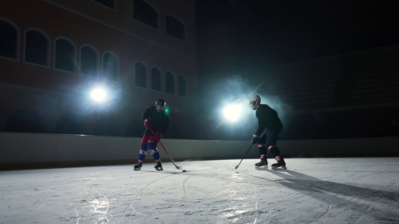 两名运动员曲棍球运动员在冰场上滑行和运球，用棍子打冰球。穿着制服、戴着头盔、拿着棍棒的年轻人在灯光昏暗的溜冰场里滑冰。慢动作视频素材