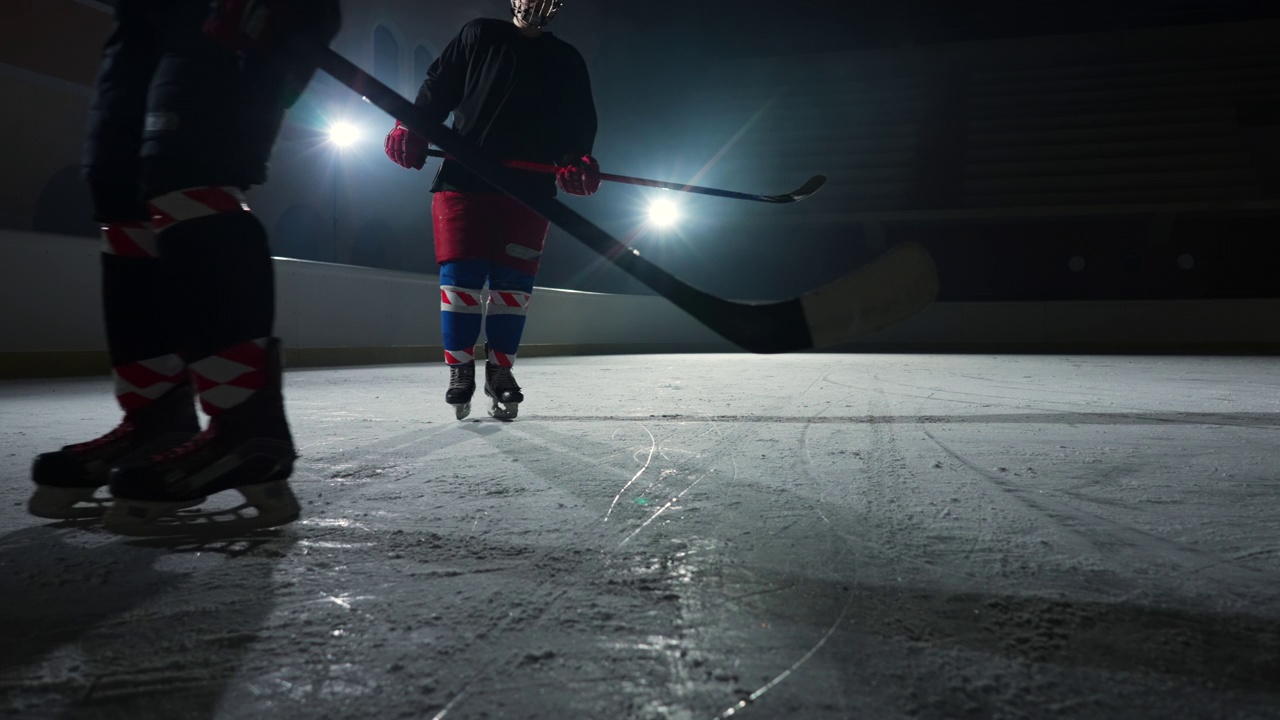 两名运动员曲棍球运动员在冰场上滑行和运球，用棍子打冰球。穿着制服、戴着头盔、拿着棍棒的年轻人在灯光昏暗的溜冰场里滑冰。慢动作视频素材