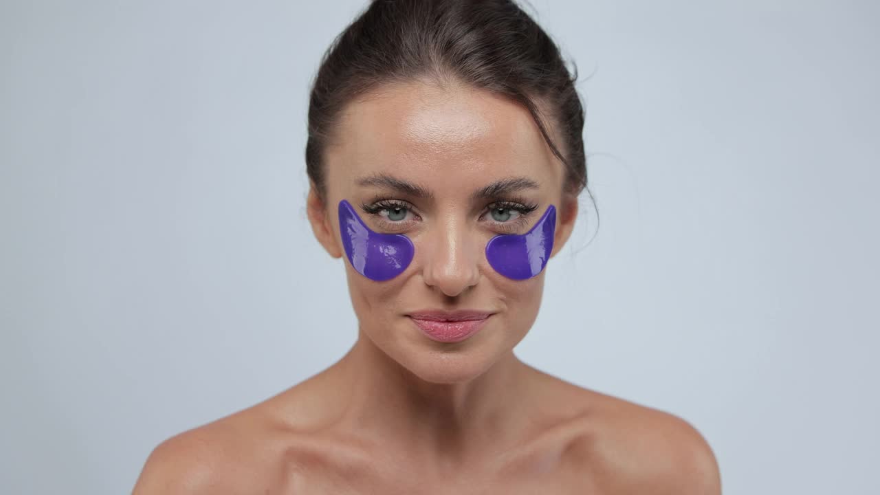 女人用紫色的眼罩滋润皮肤，对着镜头微笑，在灰色的背景上摆姿势。面部护肤美容产品理念。视频素材