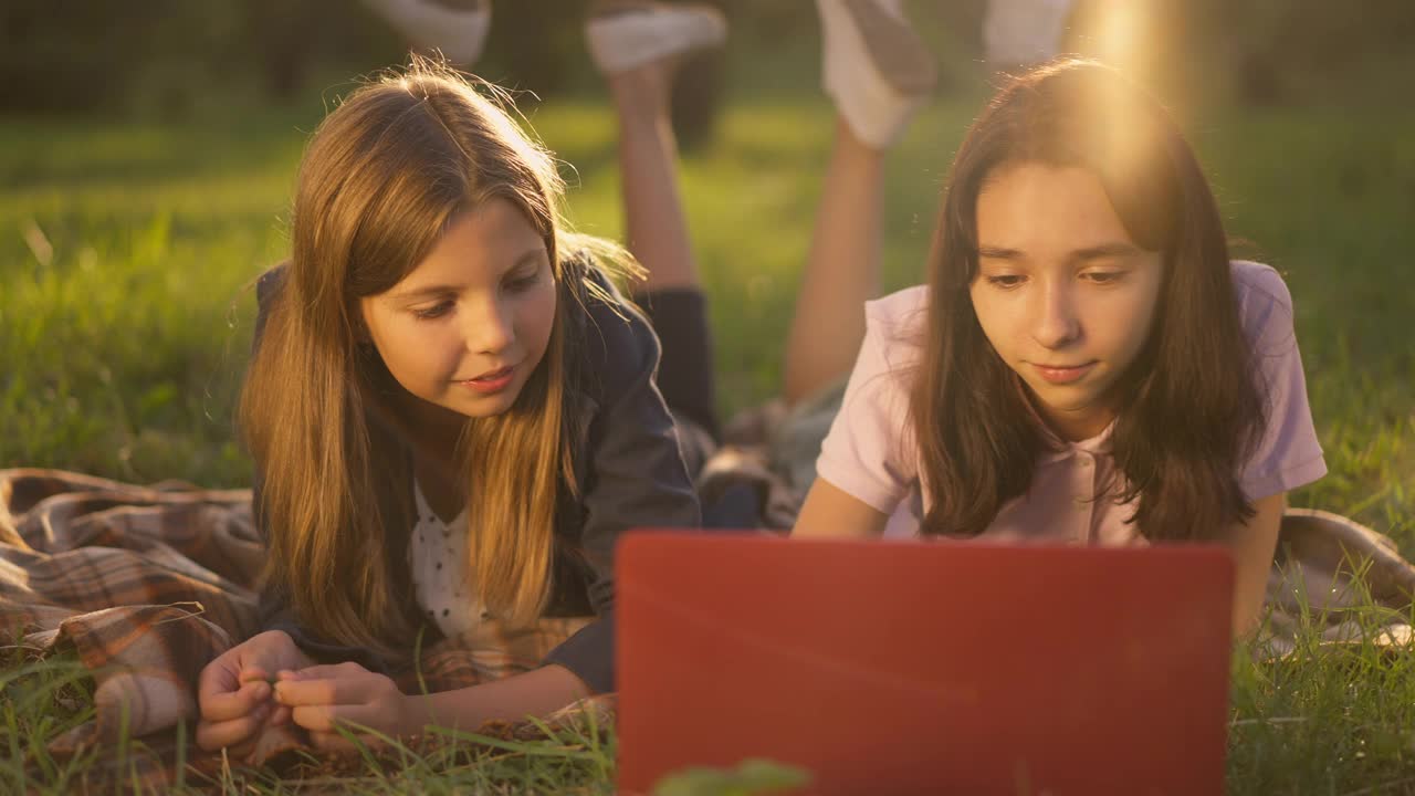 两个年轻的白人女孩躺在公园的草坪上聊天，用笔记本电脑上网。烦恼的女孩滚动眼睛的前视图肖像，朋友在慢动作说话。视频下载