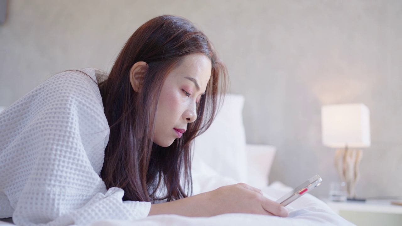 放松的亚洲女人躺在床上，用智能手机聊天视频素材
