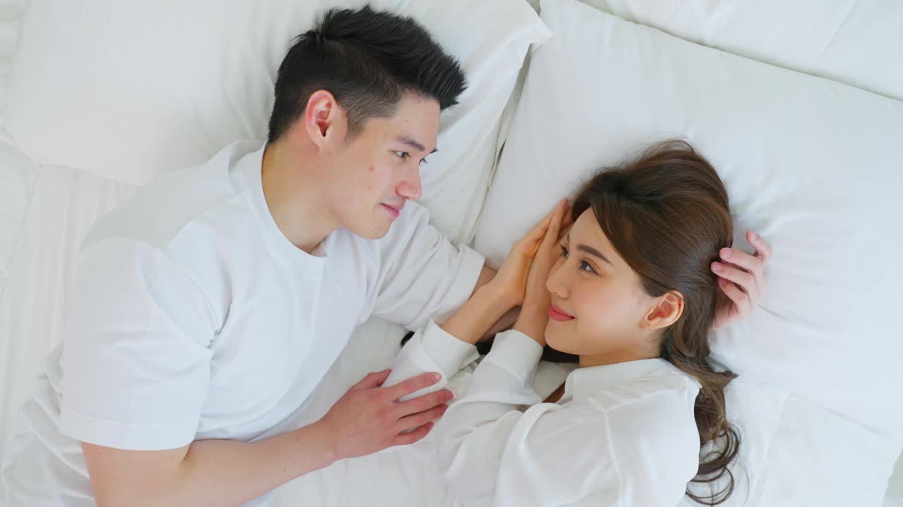 亚洲新婚夫妇躺在床上互相看对方的肖像。漂亮漂亮的年轻男女穿着睡衣在家里的卧室里享受清晨的活动。家庭关系的概念视频素材