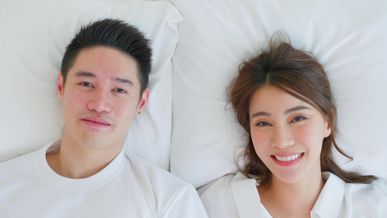 亚洲新婚夫妇躺在床上互相看对方的肖像。漂亮漂亮的年轻男女穿着睡衣在家里的卧室里享受清晨的活动。家庭关系的概念视频素材
