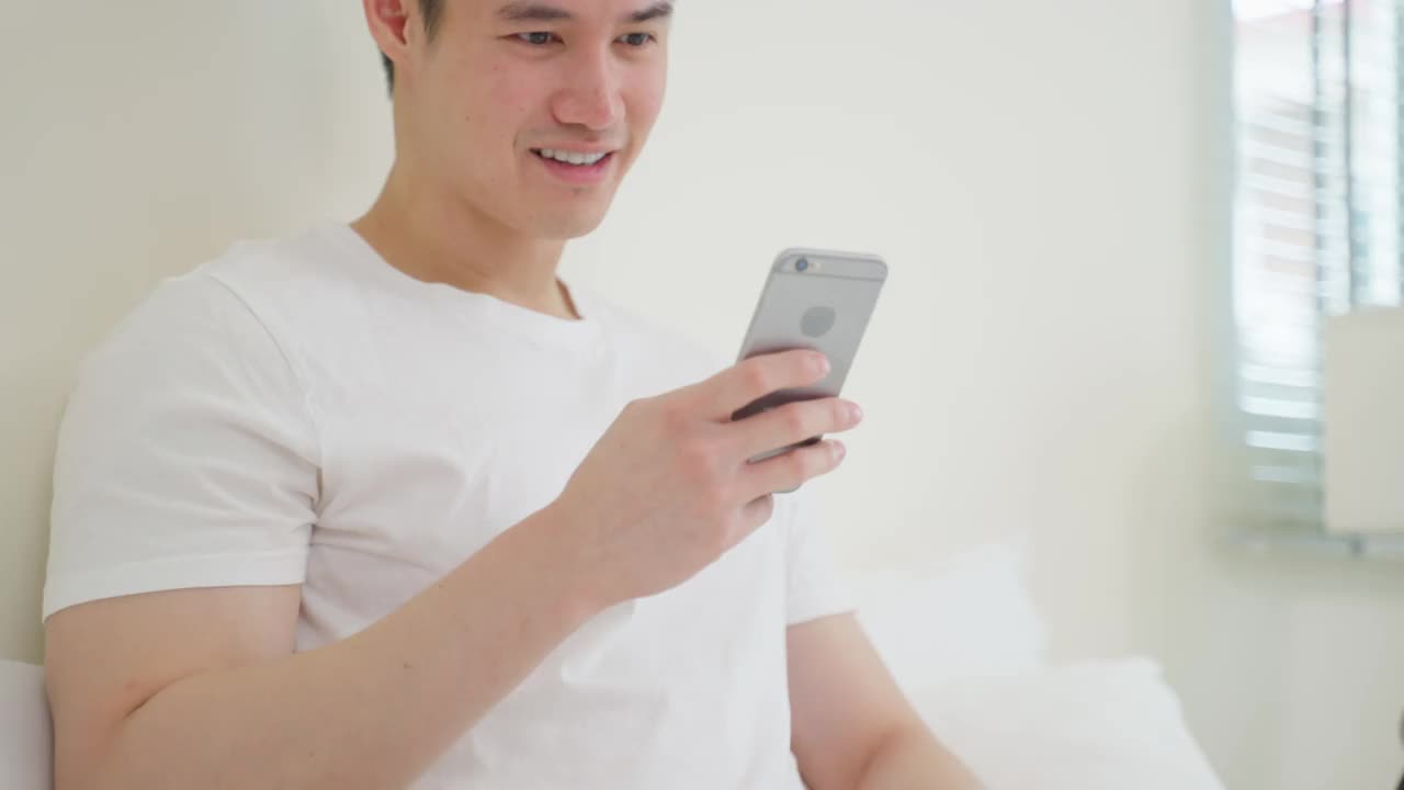 亚洲帅哥喜欢早上在家床上网购。英俊的男人躺在卧室里，拿着信用卡在家里用手机在网上商店购物后进行网上银行支付。视频素材