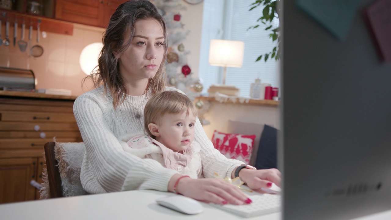 在COVID-19大流行期间，带着孩子的母亲在家里用电脑工作到很晚。视频下载