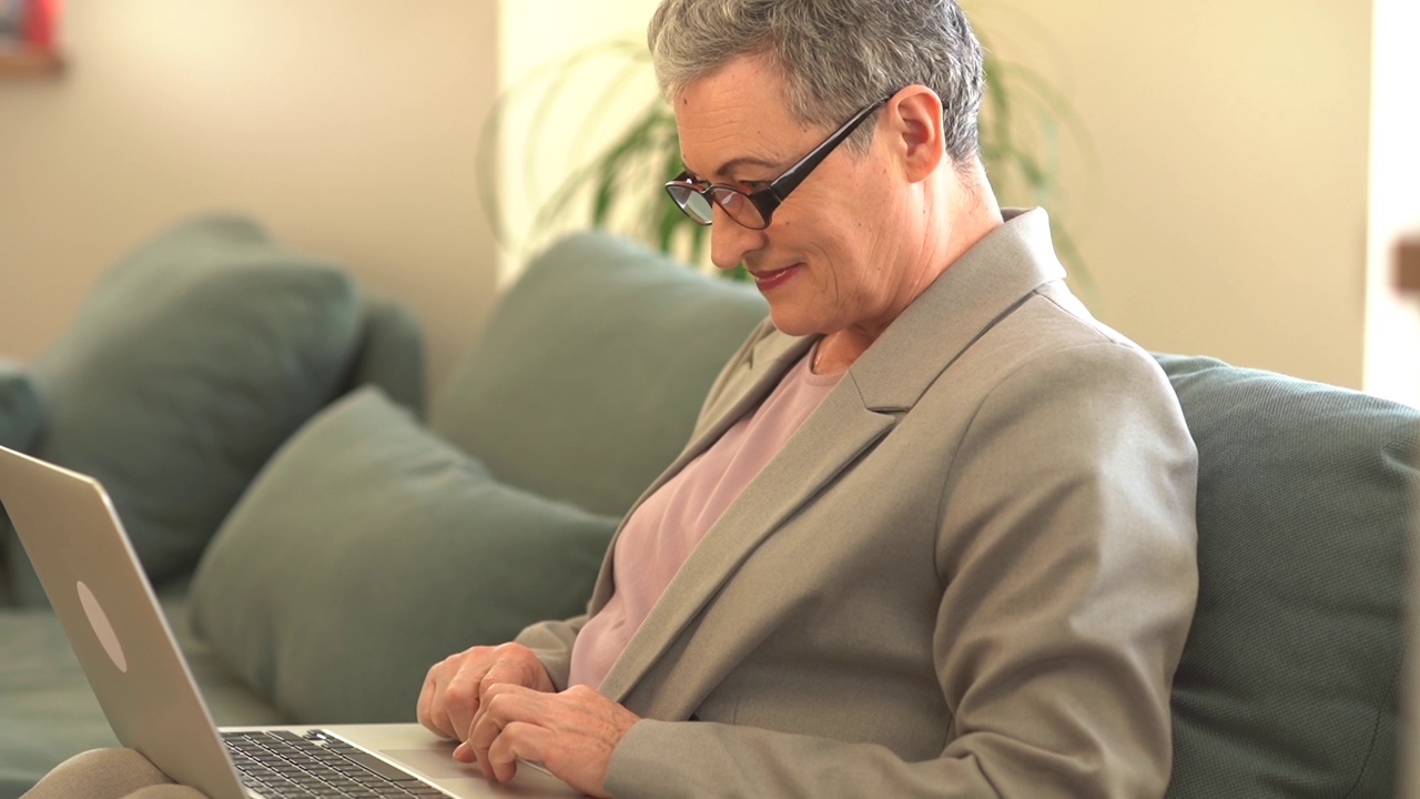 漂亮的中年商务女士戴着眼镜，穿着灰色的西装，坐在家里的沙发上用笔记本电脑工作。远程工作视频下载