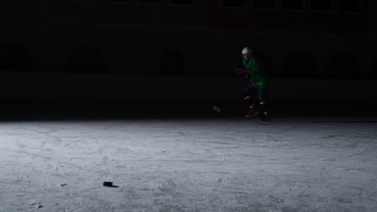 身穿绿色制服，手拿钢盔的前锋在冰面上滑行并击中冰球。一名运动员在暗光的场地上滑冰。慢动作视频素材