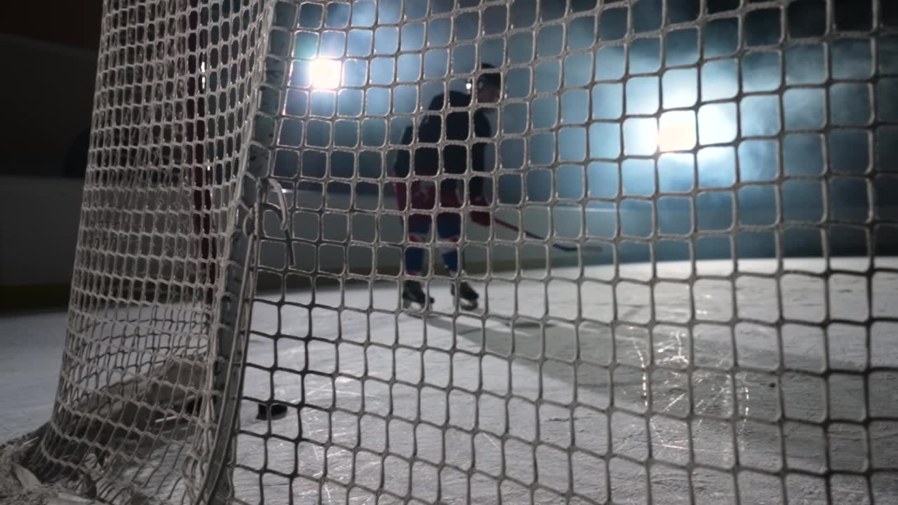 冰球前锋用球杆击中冰球并得分的画面。一个冰球飞入网的慢动作特写。有聚光灯和烟雾的黑暗冰球竞技场视频素材