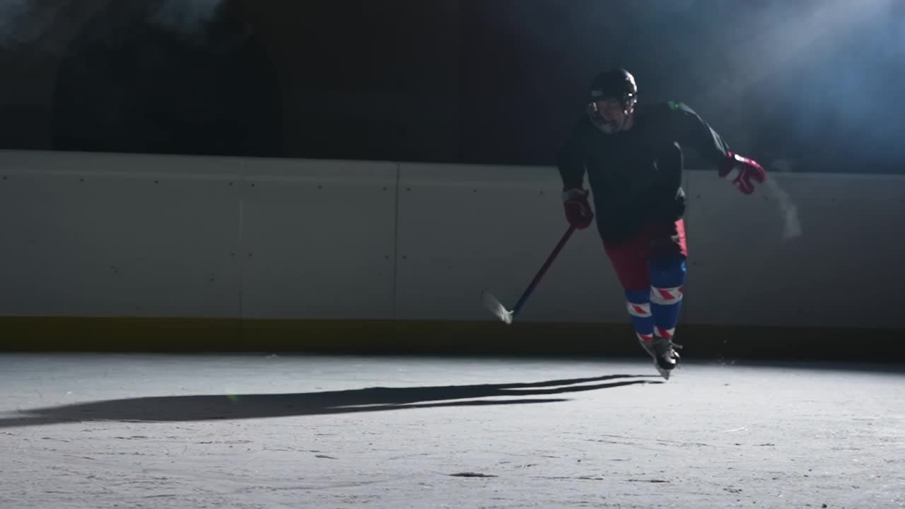 曲棍球运动员滑过一个有聚光灯和烟雾的黑暗的冰场，用曲棍球棍击中冰块，并将冰粉扔进摄像机。缓慢的运动。近距离视频素材