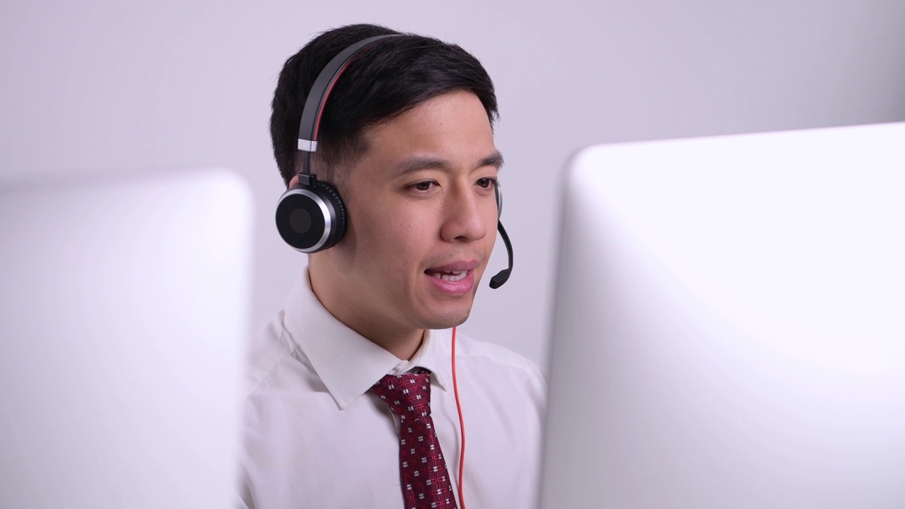 亚洲呼叫中心的接线员，面带微笑，面带麦克风，坐在办公室客户服务部的电脑前。视频购买
