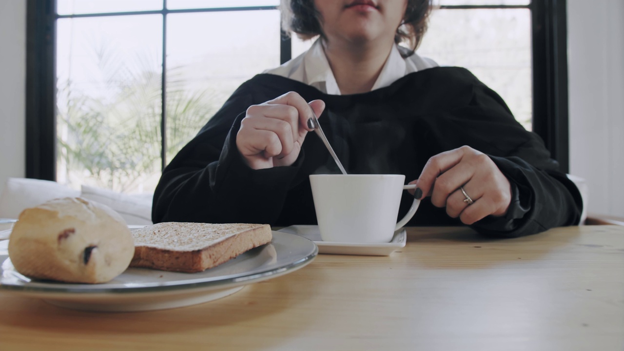 年轻女子穿着一件黑色的毛衣，悠闲地坐着，拿着勺子在家里的桌子上搅拌咖啡或茶在白色的杯子和牛奶面包视频素材