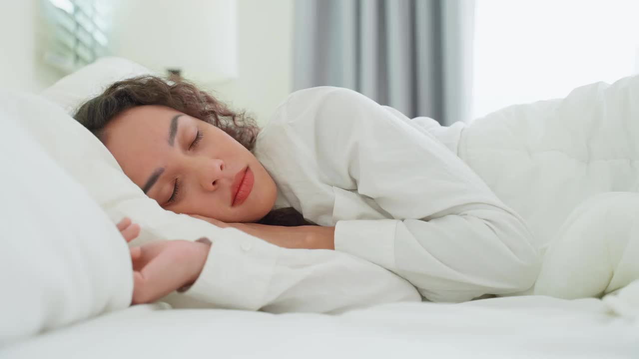 拉丁美人躺在床上的卧室在清晨。漂亮的年轻女性穿着睡衣独自睡在舒适的枕头和舒适的毯子在明亮的房间里在家保健。视频素材