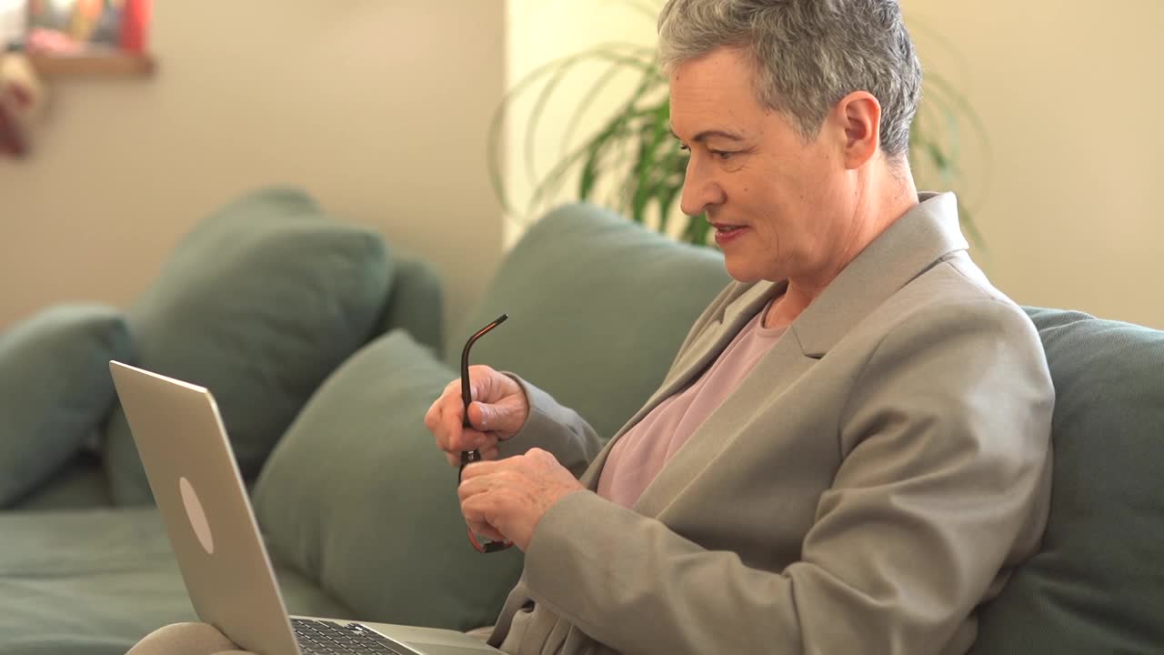一位留着灰色短发的自由职业者坐在家里的沙发上，用笔记本电脑看着摄像机视频下载